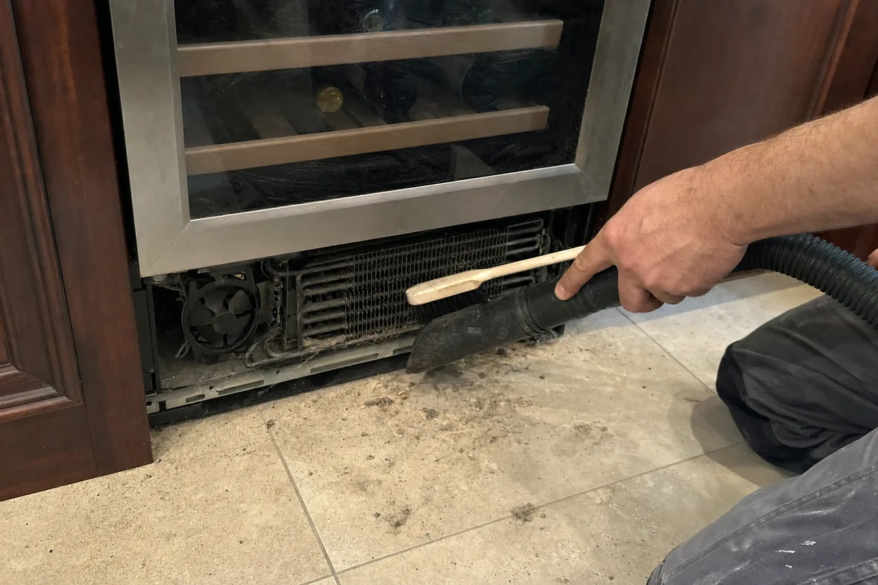 Mirrored close view of a condenser fan and fin pack behind a removed toe grille, dust drawn out onto the tile