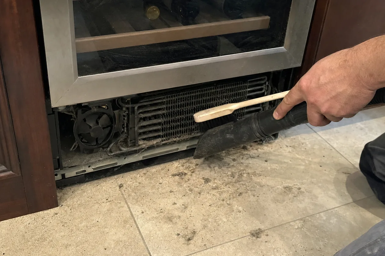 Mirrored floor-level frame of condenser cleaning at a built-in cabinet, brush in one hand and hose in the other