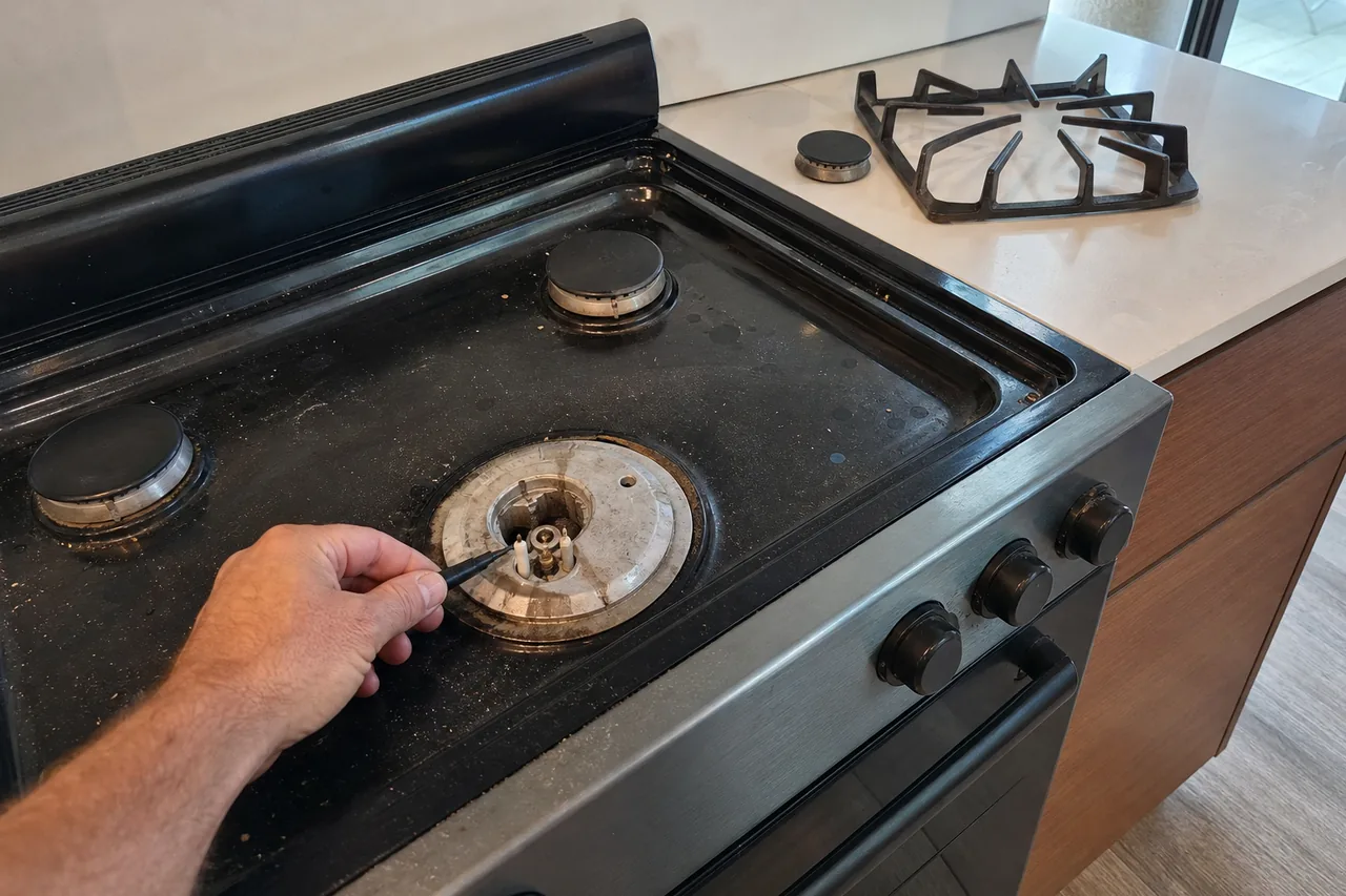 Hand guiding a thin black pick toward the igniter of an uncapped gas burner on a dark range top, its cap and grate parked on a white quartz counter beside it