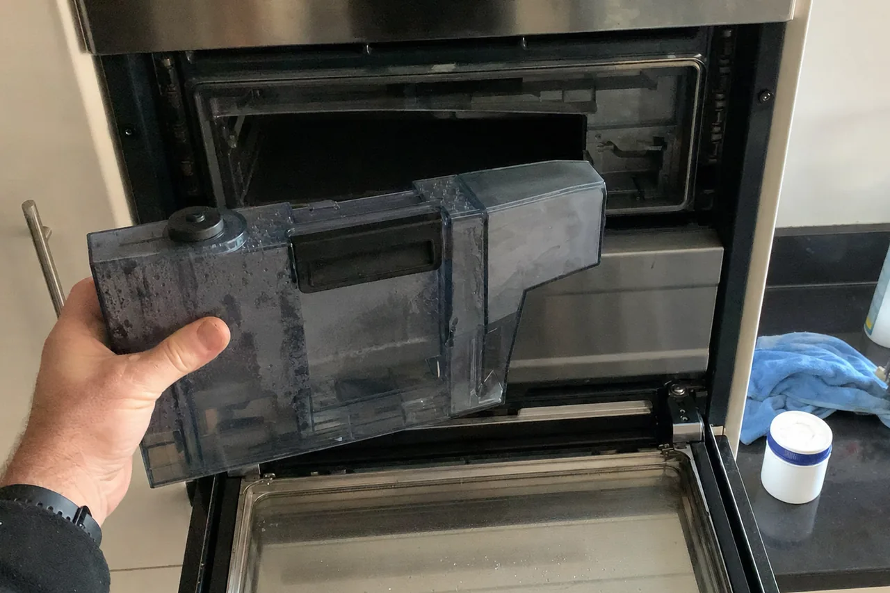 Technician holding the clouded water reservoir withdrawn from a built-in oven, the cavity open behind it
