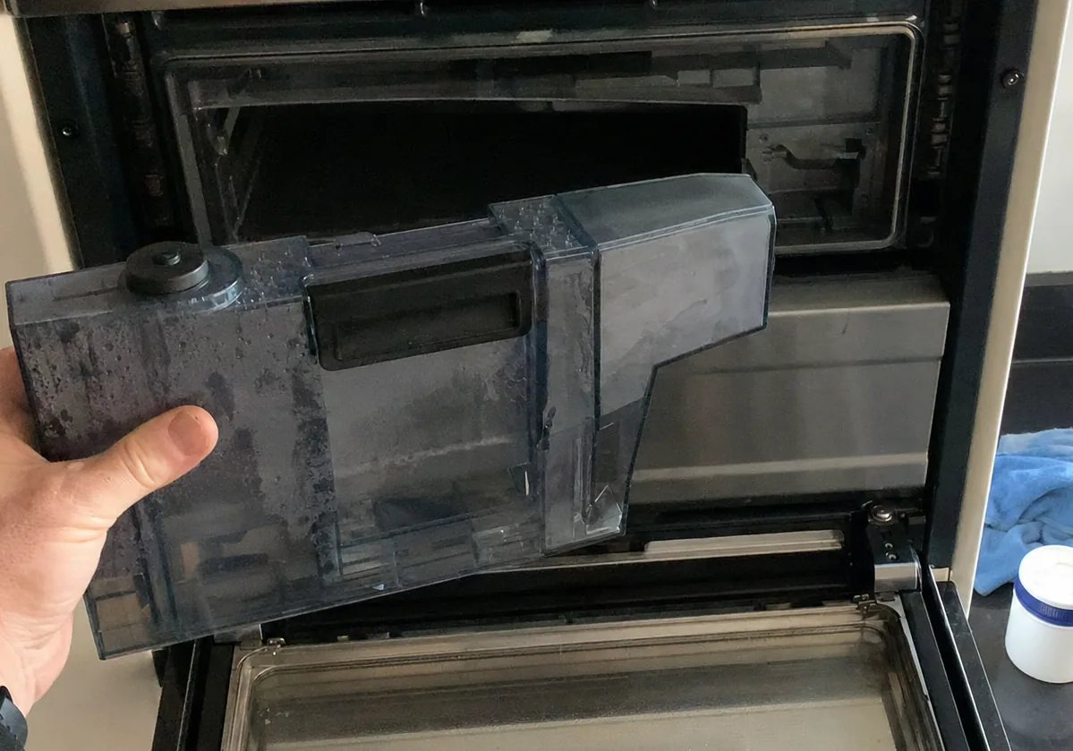 Centred frame of a plastic reservoir lifted from a stainless built-in appliance, the cavity dark behind it